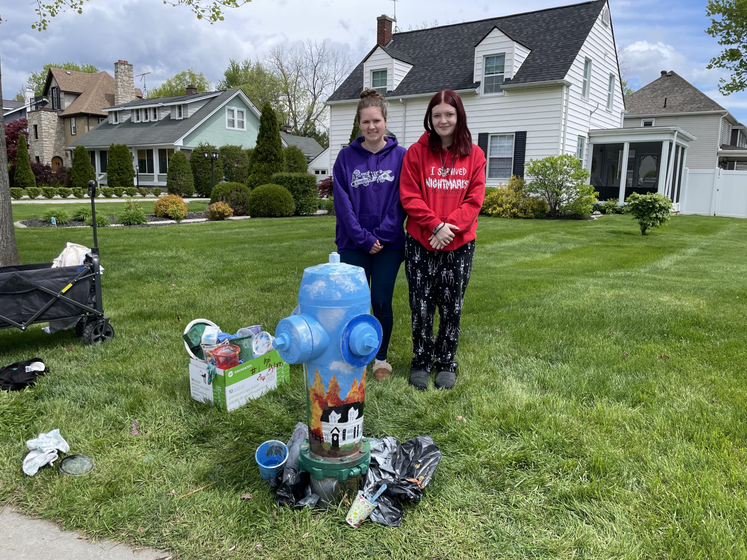 Two students post with the fire hydrant they painted. Art supplies and the house they painted on the hydrant are in the background