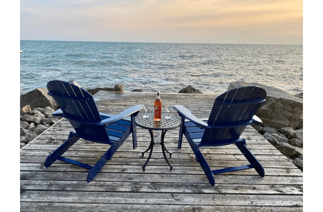 Adirondack chairs on a dock, facing the water, with a bottle of wine and two wine glasses.