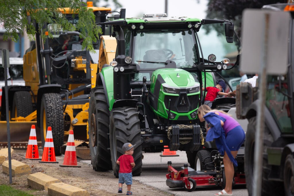 side of a street with two big tractors on it, family of mom and two young boys playing on the closest tractor