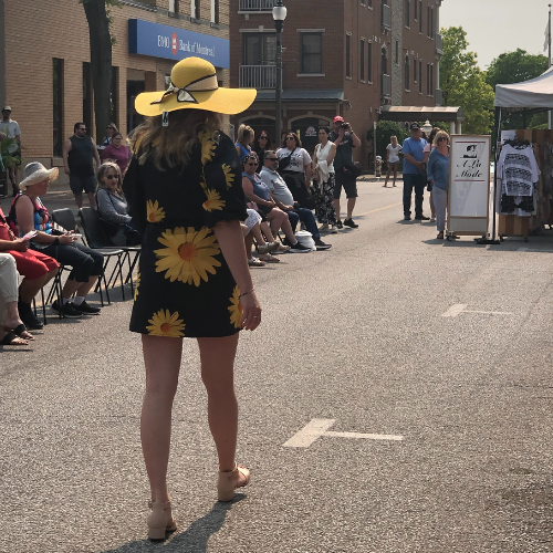 girl in sunflower dress with big yellow hat walking down the street, peopled lined up in chairs on the side, like a fashion show