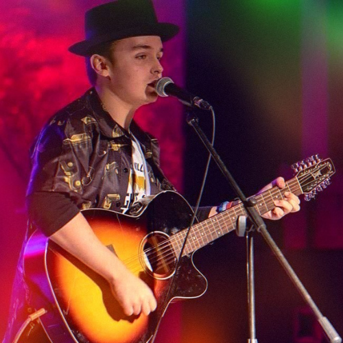 guy (Justin Jewell) with a fedora hat playing the guitar singing into a mic on stage