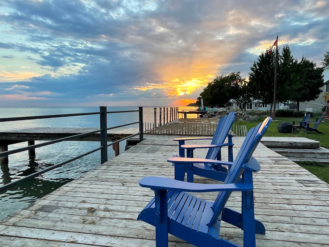 Blue muskoka chairs on a dock overlooking the lake with a sunset in the background